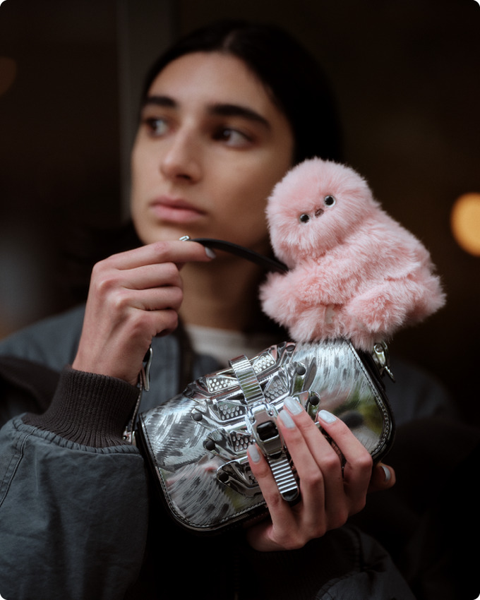 A pink fluffy plush creature with a round body and small black eyes sits atop a silver metallic shoulder bag with decorative metal hardware and straps, held by a person wearing dark clothing.