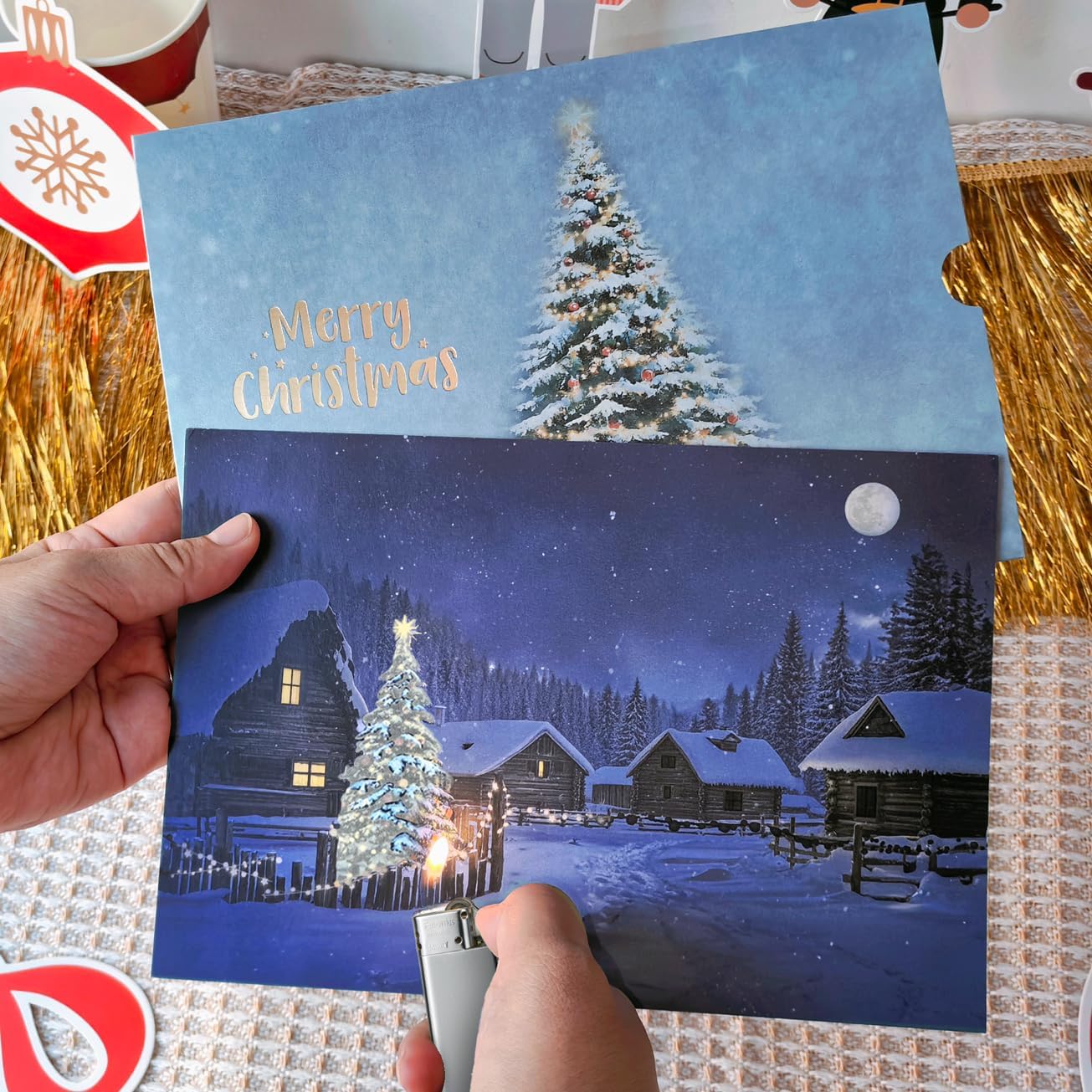 A festive greeting card featuring dual holiday scenes that change when warmed: the top shows a snow-covered Christmas tree against a light blue sky, while the bottom reveals a magical winter village with illuminated log cabins, a decorated tree, and moonlit forest landscape in deep blue tones.
