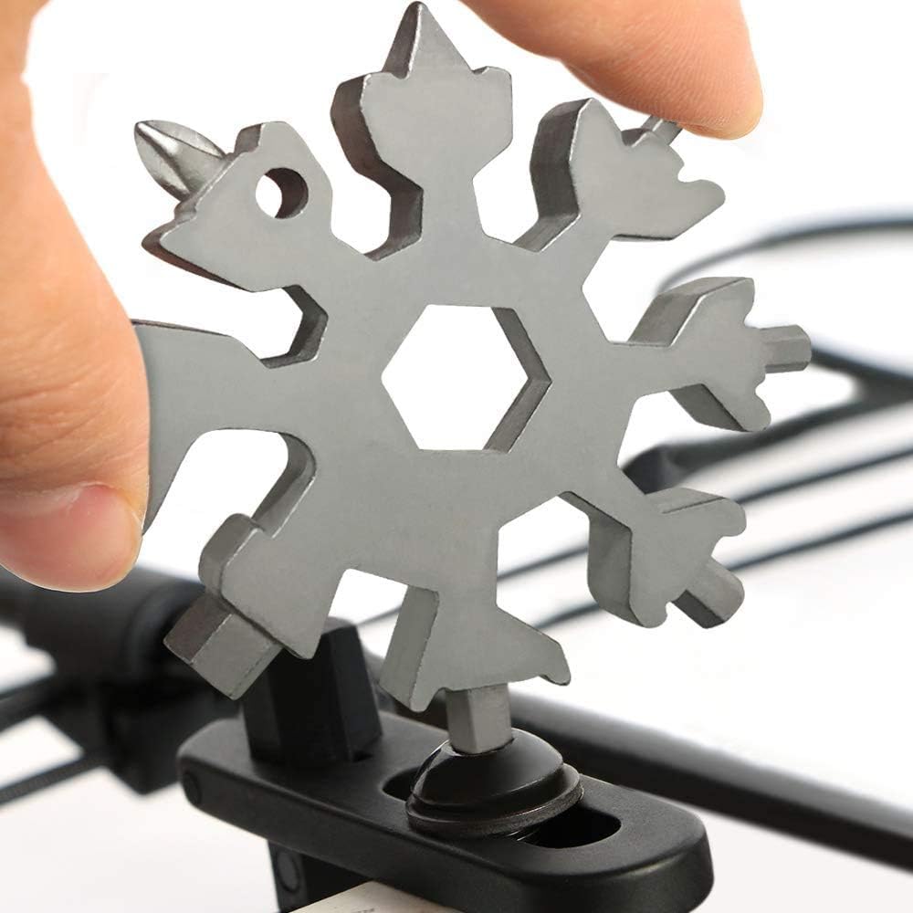 A stainless steel snowflake-shaped multi-tool held in someone's hand, featuring various integrated tools including a hexagonal wrench, screwdrivers, and other utility implements around its perimeter, mounted on a black rubber base.