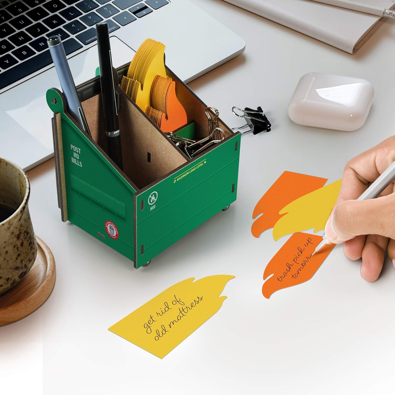 A green miniature dumpster-shaped desk organizer holding colorful tape dispensers and writing supplies, paired with matching flame-shaped sticky notes in yellow and orange with humorous handwritten text on a workspace desk.