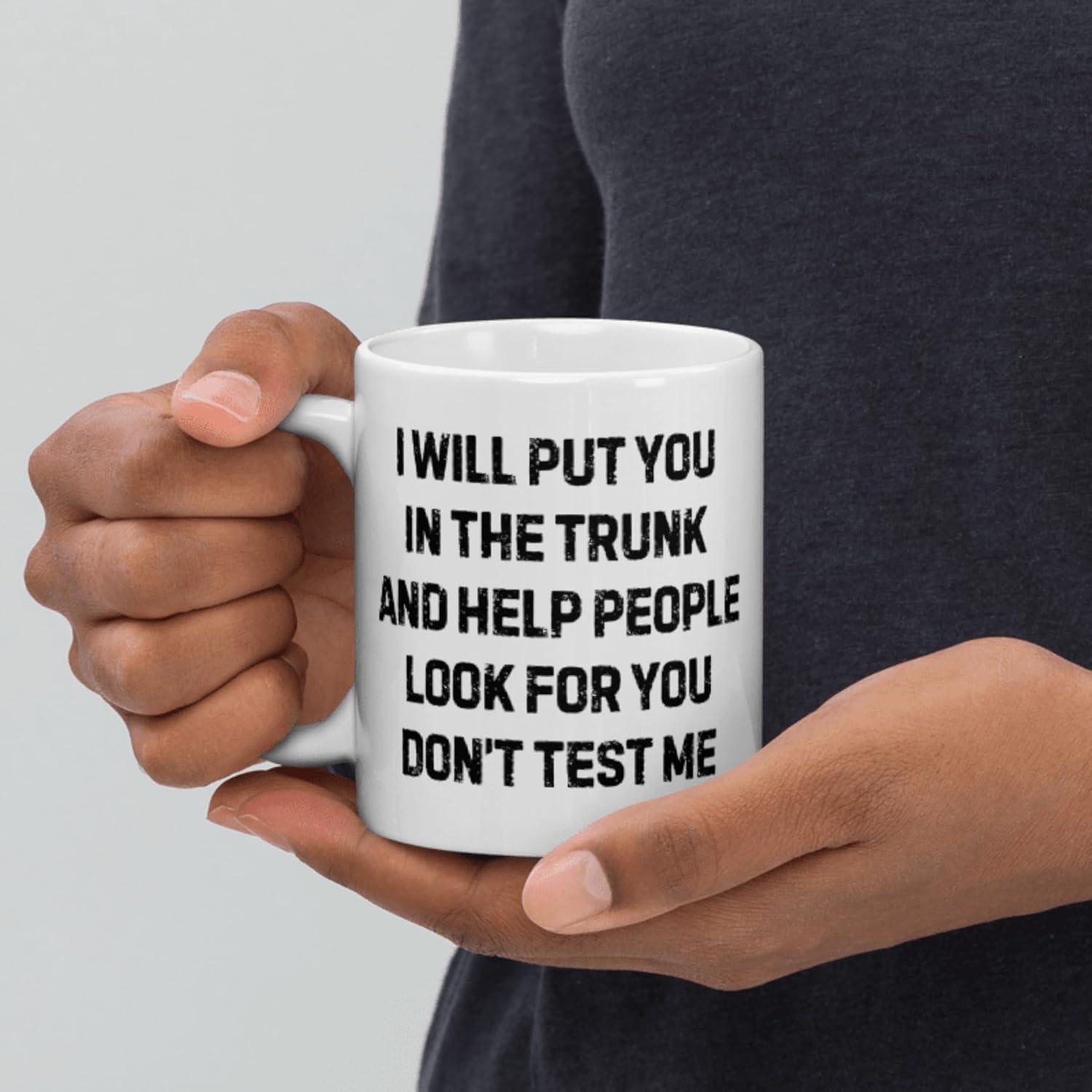 A white ceramic mug held in someone's hands features black text reading "I WILL PUT YOU IN THE TRUNK AND HELP PEOPLE LOOK FOR YOU DON'T TEST ME" as a humorous warning message.