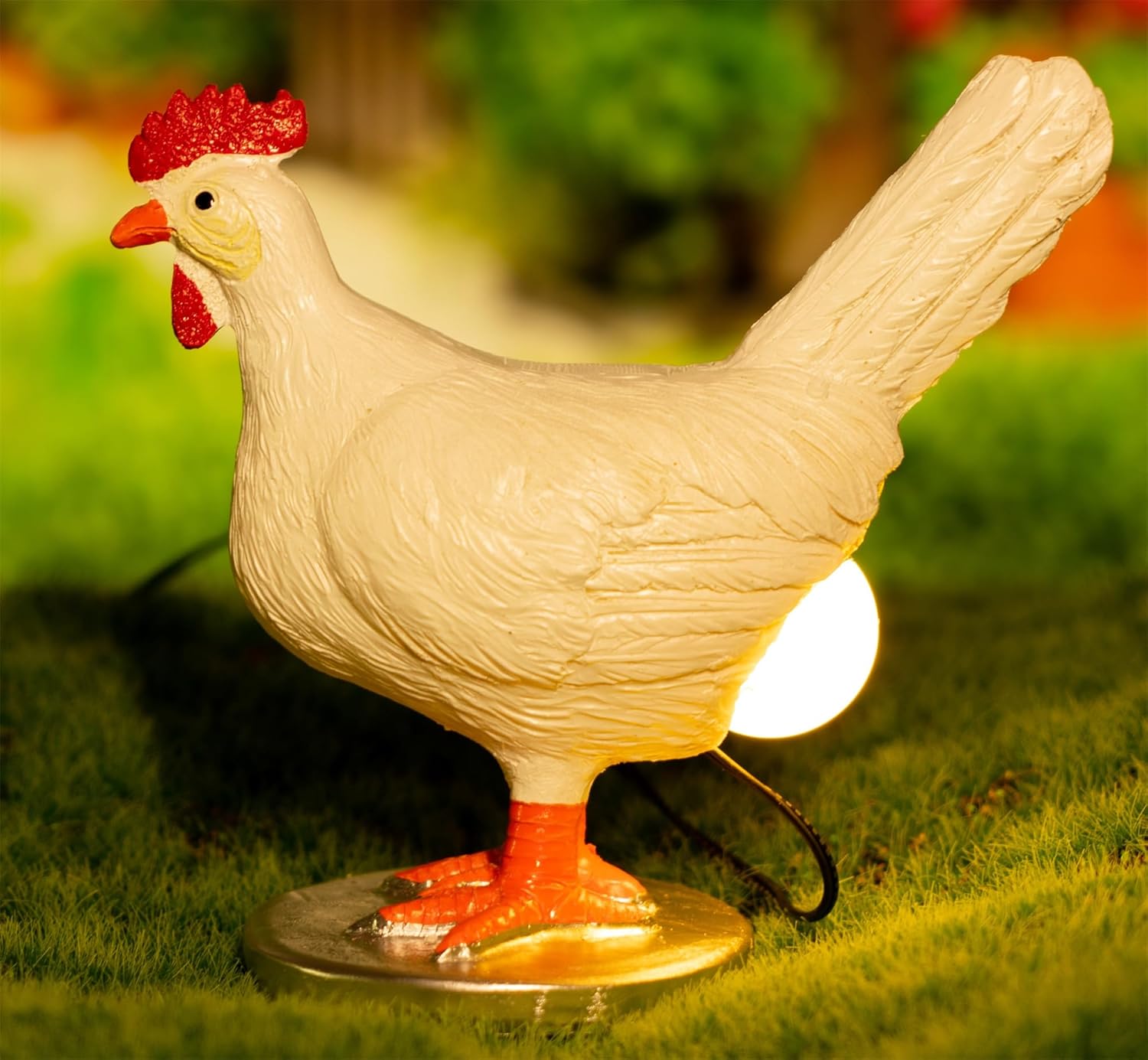 A whimsical cream-colored chicken night light with a bright red comb and orange beak, featuring an illuminated white belly that glows softly, displayed on grass with a blurred garden background.