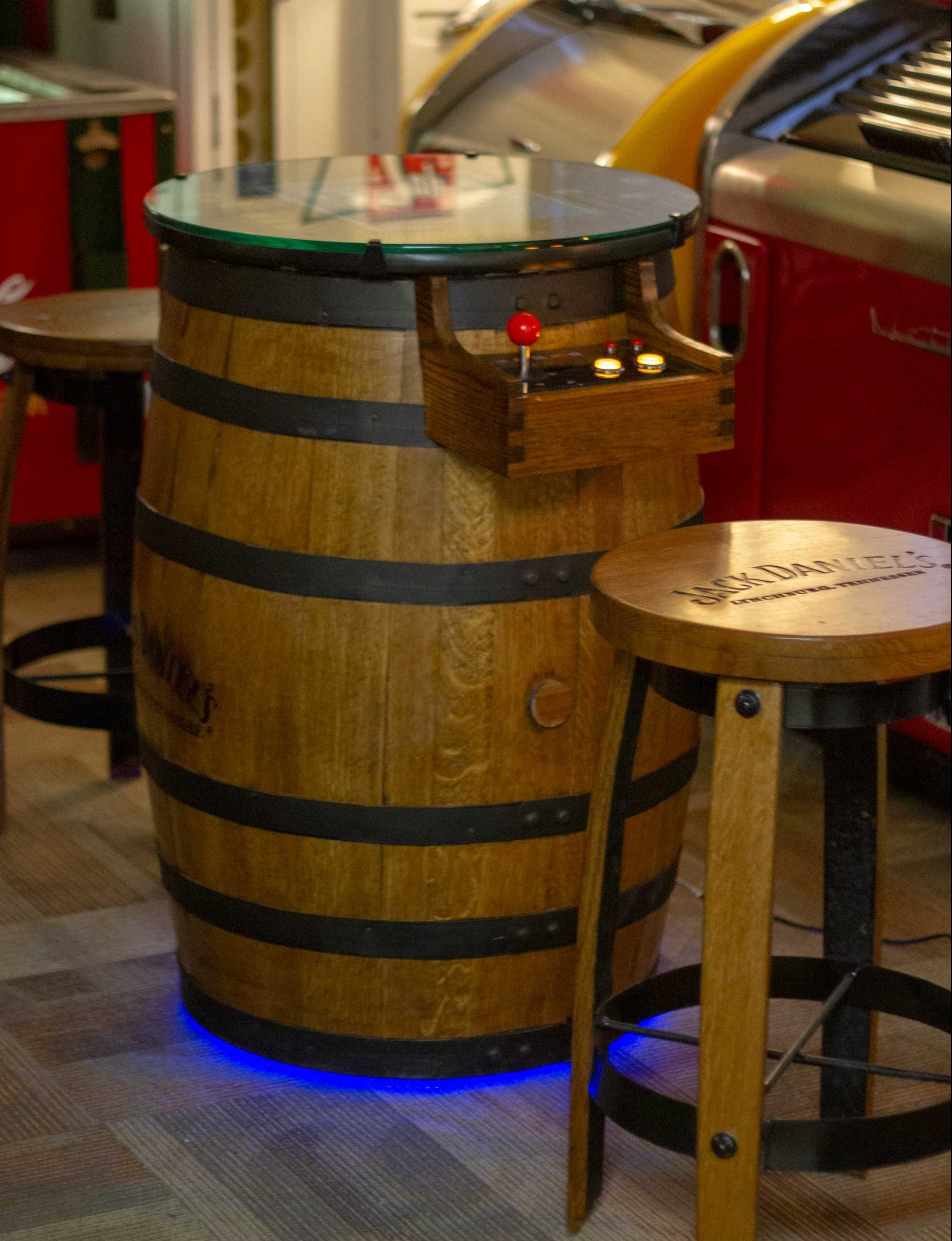 for Whiskey Barrel Arcades

A wooden whiskey barrel with metal bands serves as an arcade game cabinet, featuring a glass top with integrated controls, a red joystick, and illuminated blue accent lighting at the base, accompanied by a matching wooden stool.