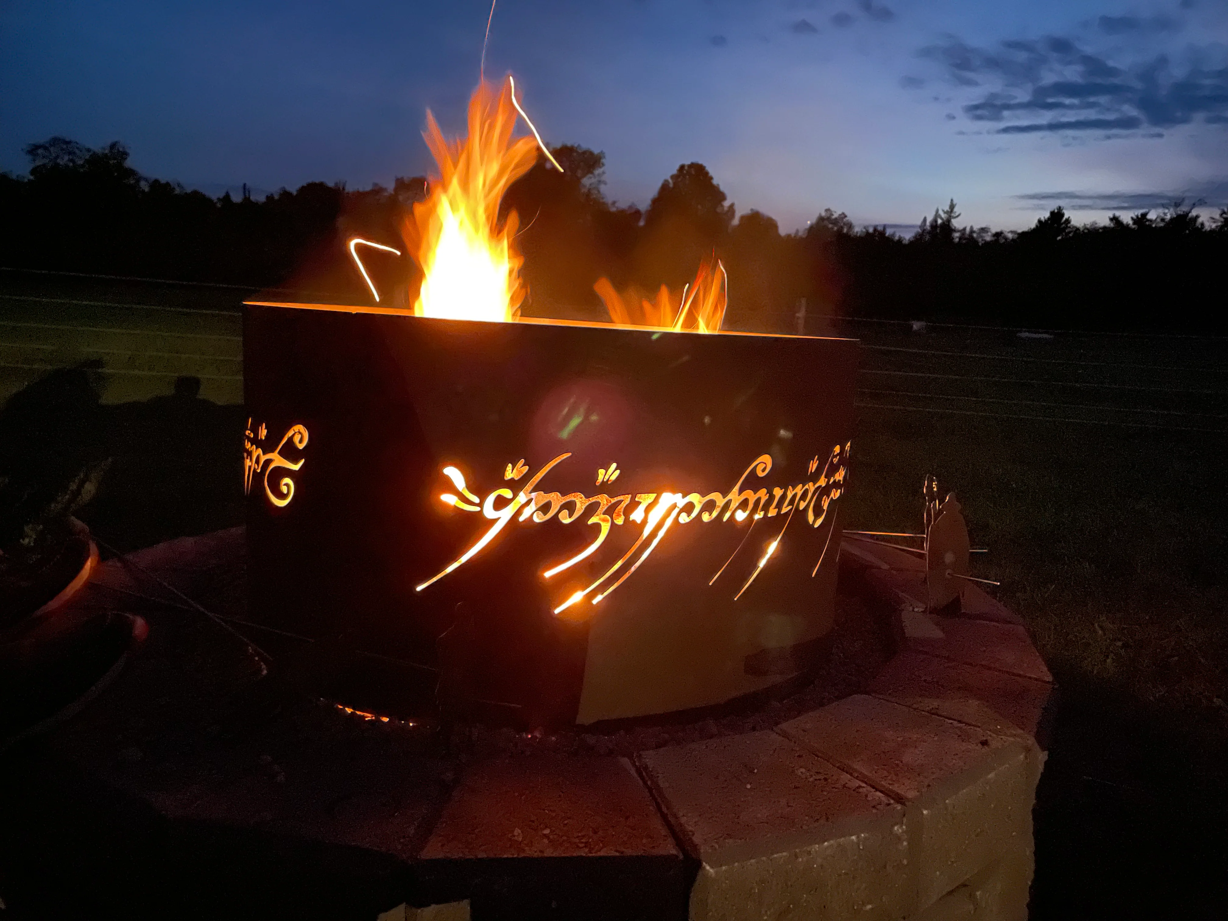 A large metal fire pit glowing with bright flames at dusk, featuring illuminated Elvish script from "The Lord of the Rings" etched across its dark steel panels.
