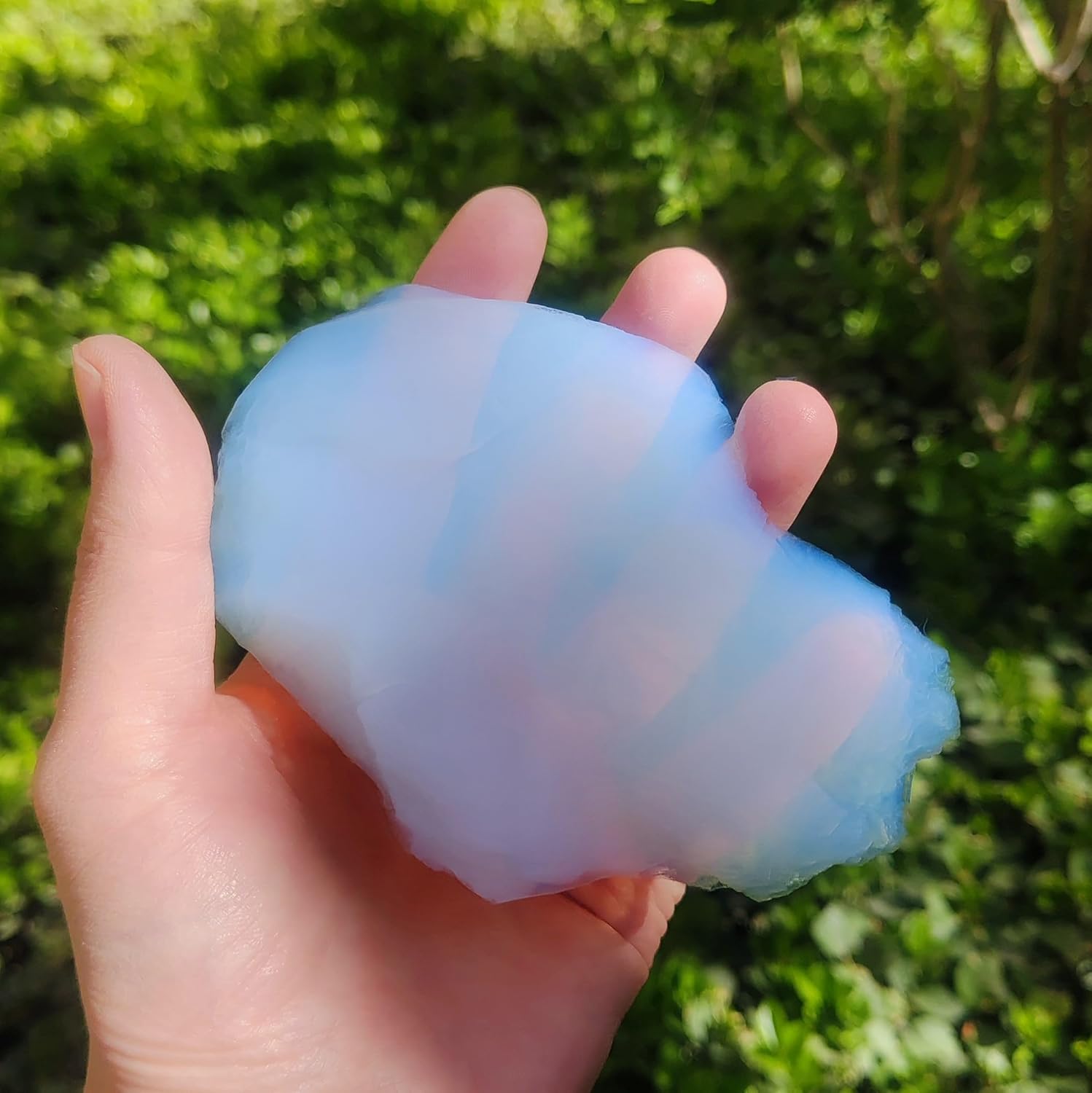 A translucent, pale blue aerogel sphere held in an open palm against a blurred green foliage background, demonstrating the extremely lightweight and airy nature of this porous solid material.