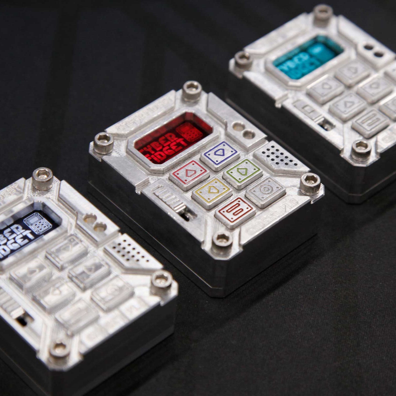 Three white modular fidget devices with customizable buttons and displays arranged on a black surface. Each unit features a small screen, mechanical switches with colored icons (red, blue, yellow, green), and tactile elements for interactive fidgeting and programming.