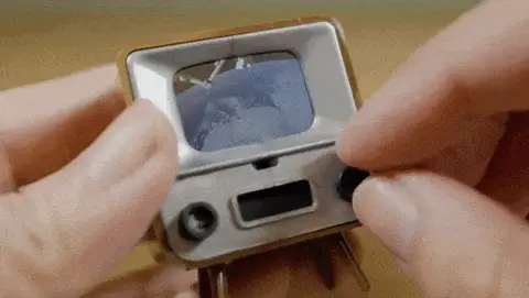 Hands holding a miniature silver television toy with a small blue screen, tiny speaker grille, and control buttons, representing a nostalgic pocket-sized recreation of a classic TV set.