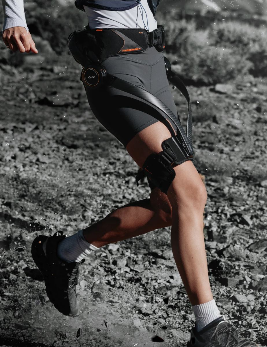 A runner wearing a black and gray Hypershell Wearable Exoskeleton leg brace with orange accents, featured on a trail with rocky terrain visible underfoot.