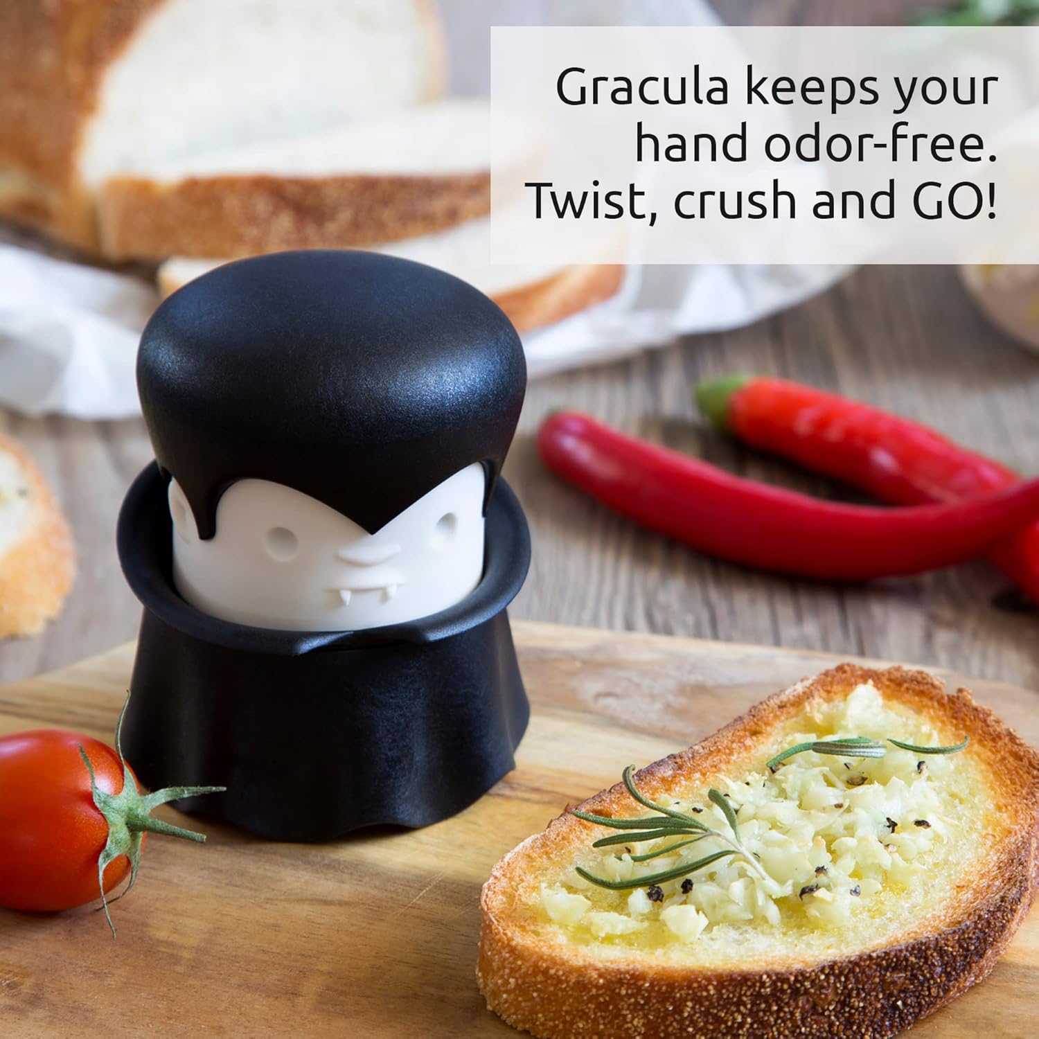 Gracula Garlic Crusher: A black ninja-shaped garlic press with a white smiling face displayed on a wooden cutting board next to fresh garlic bread, tomatoes, and red peppers, with text stating "Gracula keeps your hand odor-free. Twist, crush and GO!"