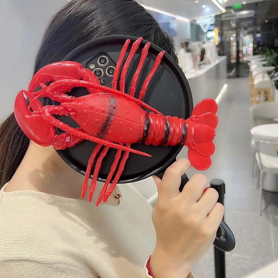 A playful red lobster-shaped iPhone case featuring long antennae and claws, displayed against a black frying pan-styled phone backing, held by a person in a retail setting.