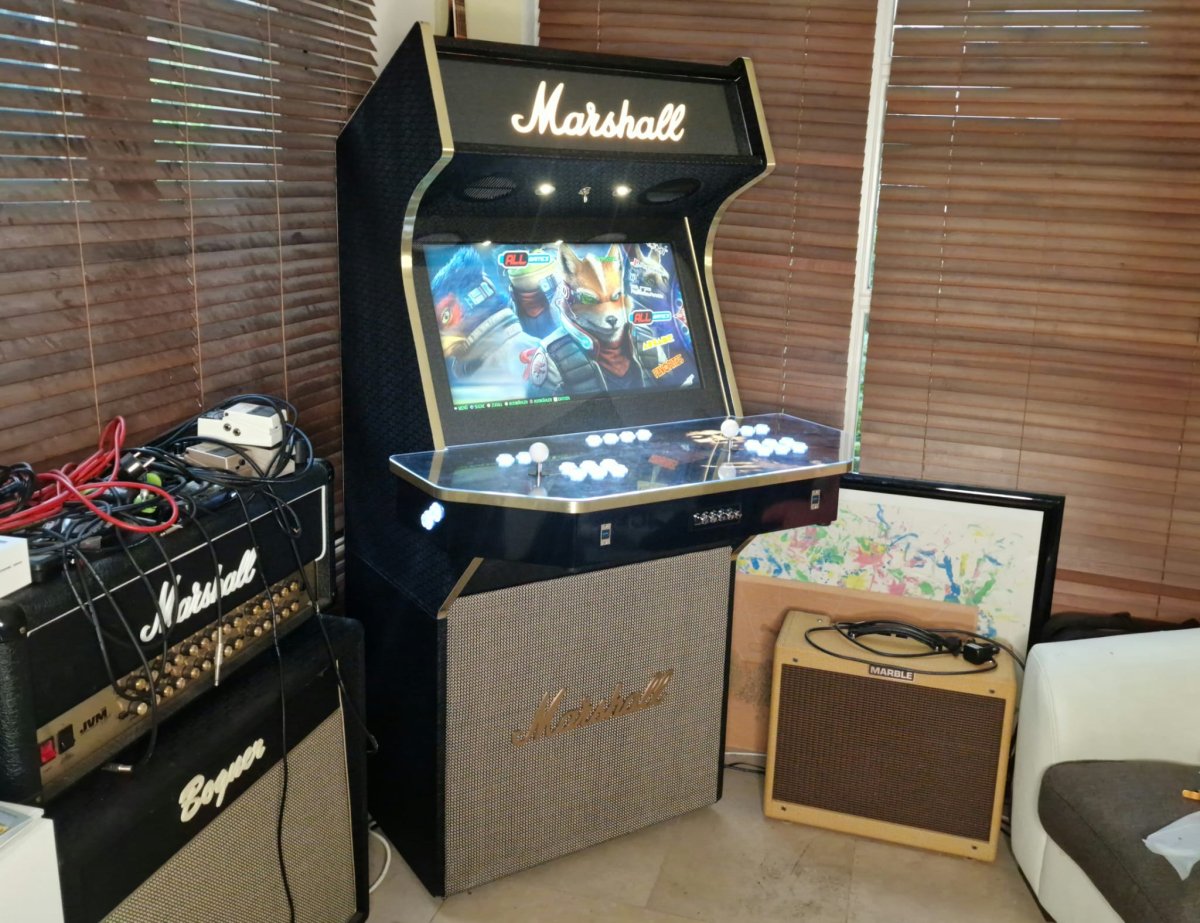 A custom arcade cabinet featuring a Marshall amplifier design, displaying a vintage fighting game on its screen with illuminated white arcade buttons on the control panel, flanked by a Marshall speaker cabinet and smaller Marshall combo amplifier in a home entertainment setup.