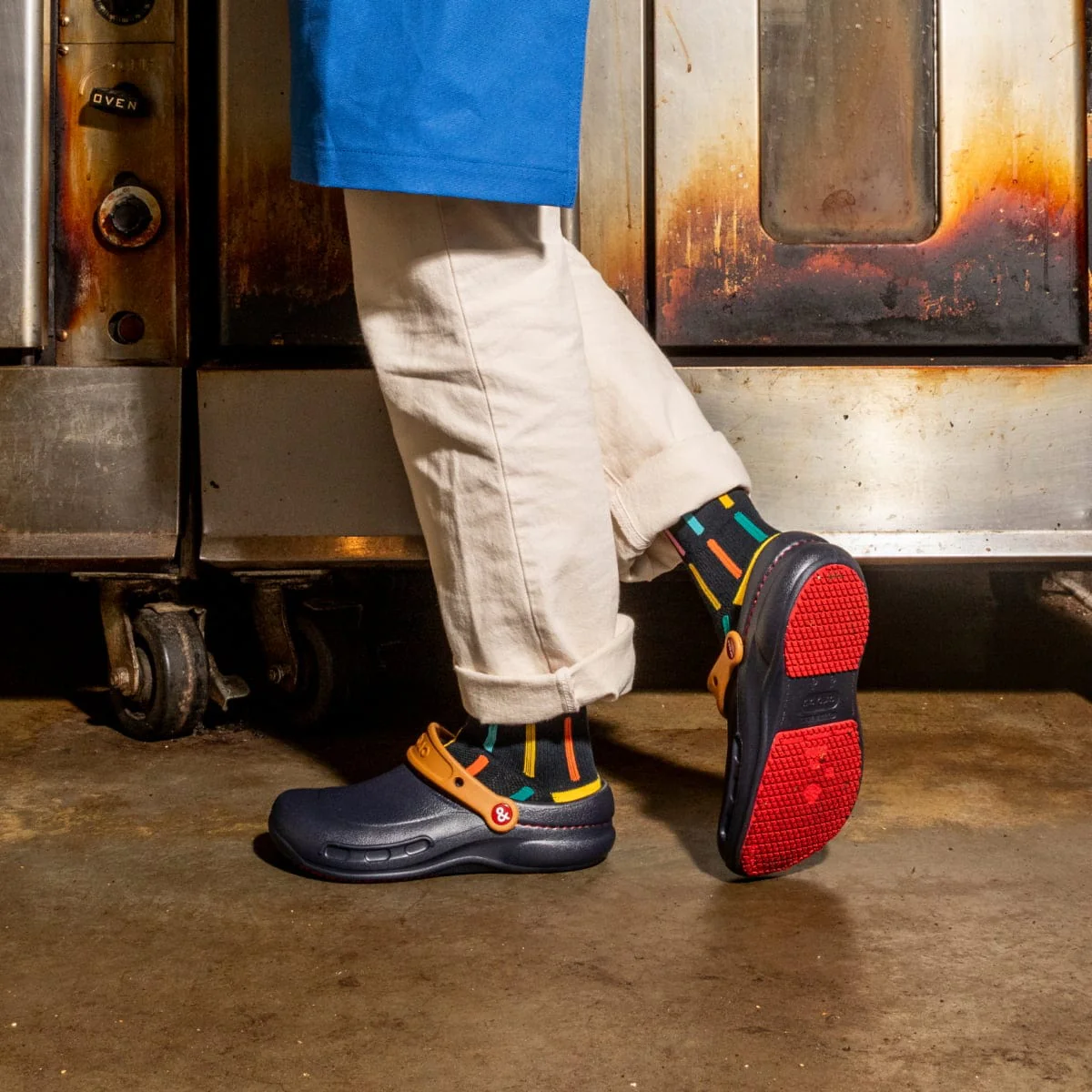 Navy blue and orange Crocs clogs with colorful striped socks, featuring red outsoles, worn by a person in white chef's pants standing in an industrial kitchen setting.
