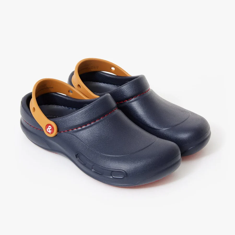Pair of navy blue clogs with tan/caramel-colored heel straps and red stitching detail, featuring branded buttons and a professional kitchen-style design from the Crocs X Hedley & Bennett collaboration.