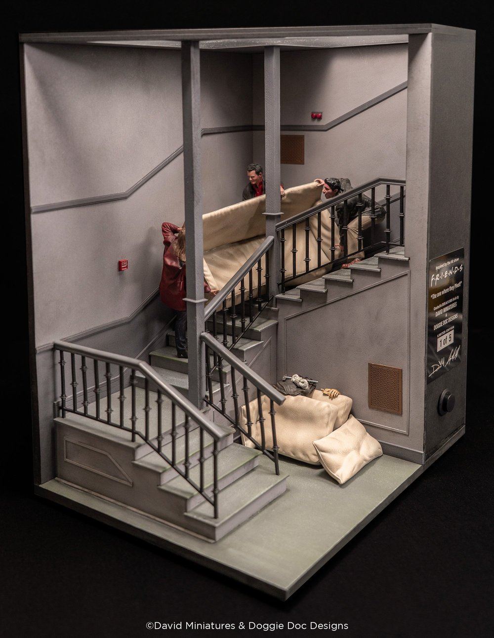 A detailed dollhouse-scale diorama recreating the iconic stairwell pivot scene from the TV show "Friends," featuring miniature figures, a beige couch on stairs, metal railings, and multi-level concrete interior architecture with realistic lighting and shadows.