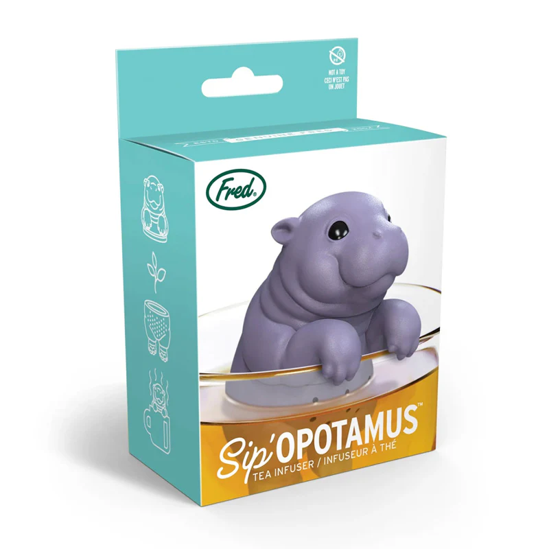 A purple silicone baby hippo-shaped tea infuser by Fred, displayed in its turquoise and yellow retail packaging. The cute hippo figurine sits on a gray leaf-shaped base with a gold-colored handle for steeping tea.